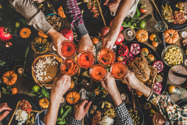 Hands raising glasses in a toast over a festive table filled with a roasted turkey, pumpkin pie, seasonal fruits, vegetables, and decorative pumpkins, evoking a warm holiday gathering.